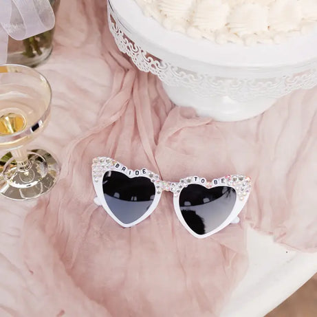 Heart-shaped sunglasses with 'Bride to Be' text on a pink fabric background.