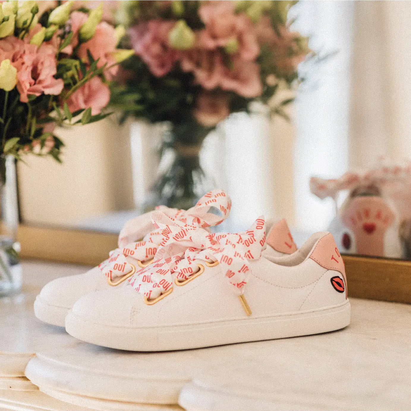 A pair of white bridal trainers with pink embroidery and silk laces placed on a surface with a bouquet of flowers in the background.