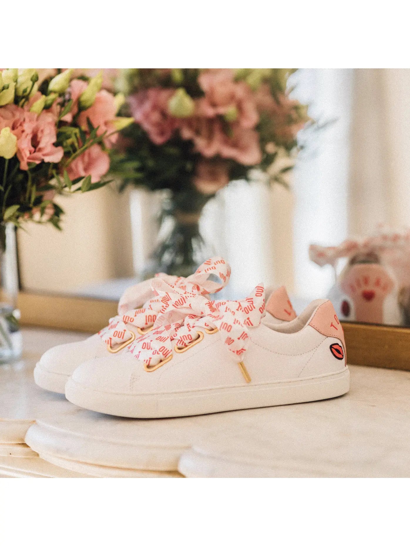 A pair of white bridal trainers with pink embroidery and silk laces placed on a surface with a bouquet of flowers in the background.