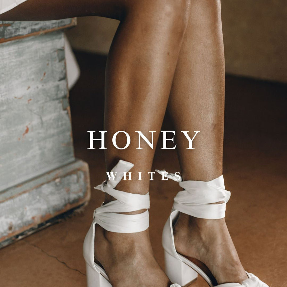 White bridal sandals with mid block heel worn by a person sitting on a wooden floor.