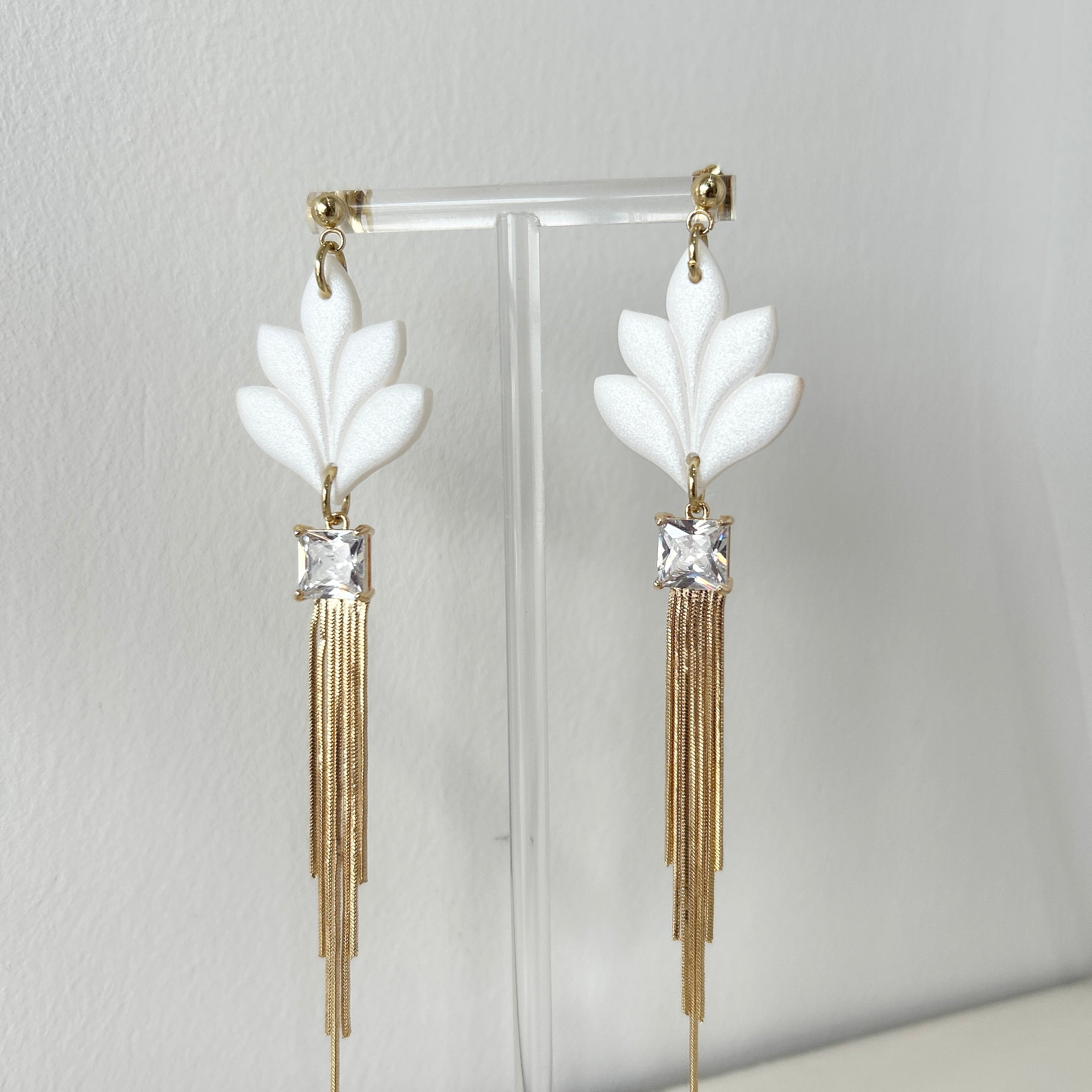Pair of statement earrings with white floral elements and gold tassels on a stand against a light gray background.
