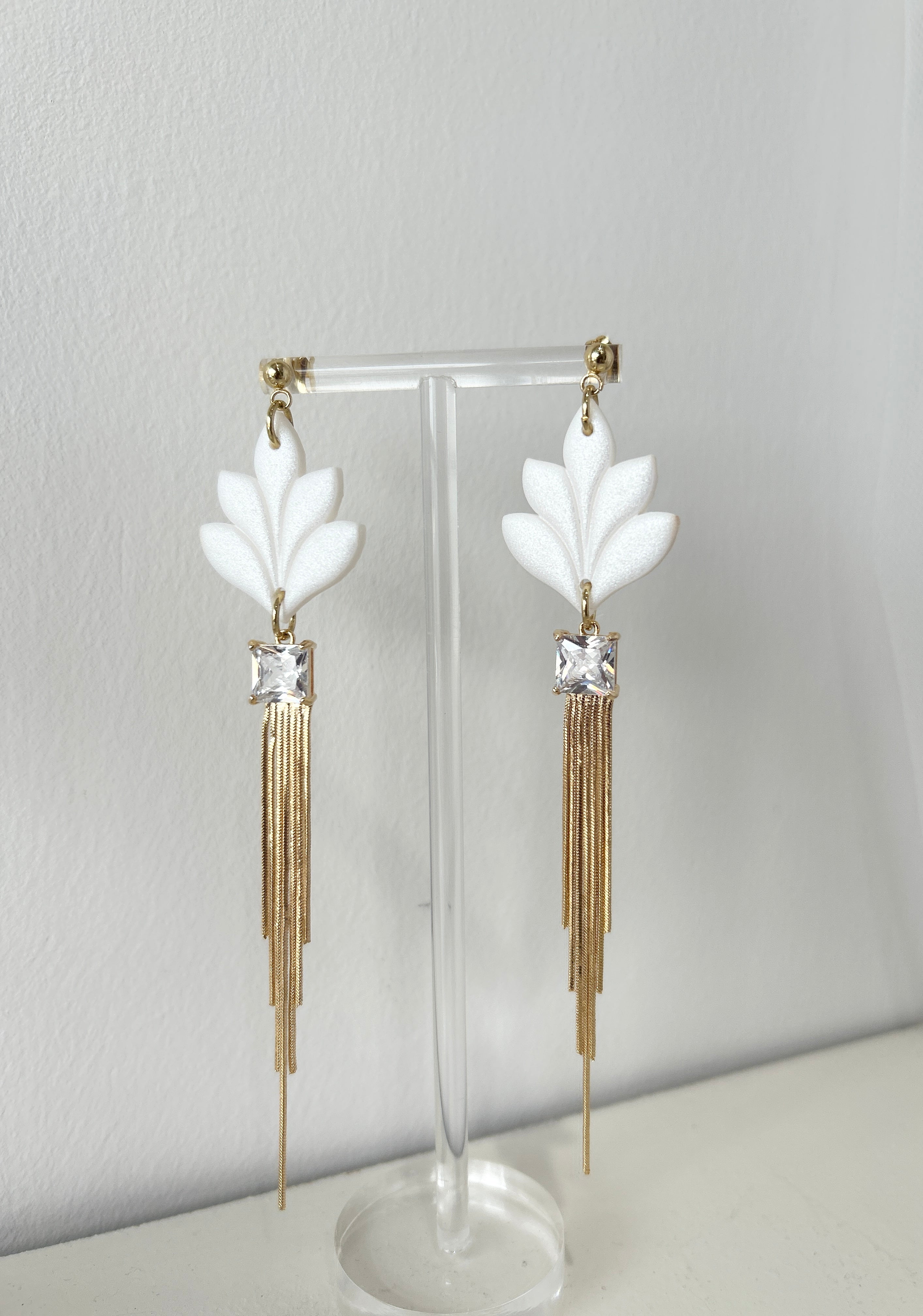 Pair of statement earrings with white floral elements and gold tassels on a stand against a light gray background.