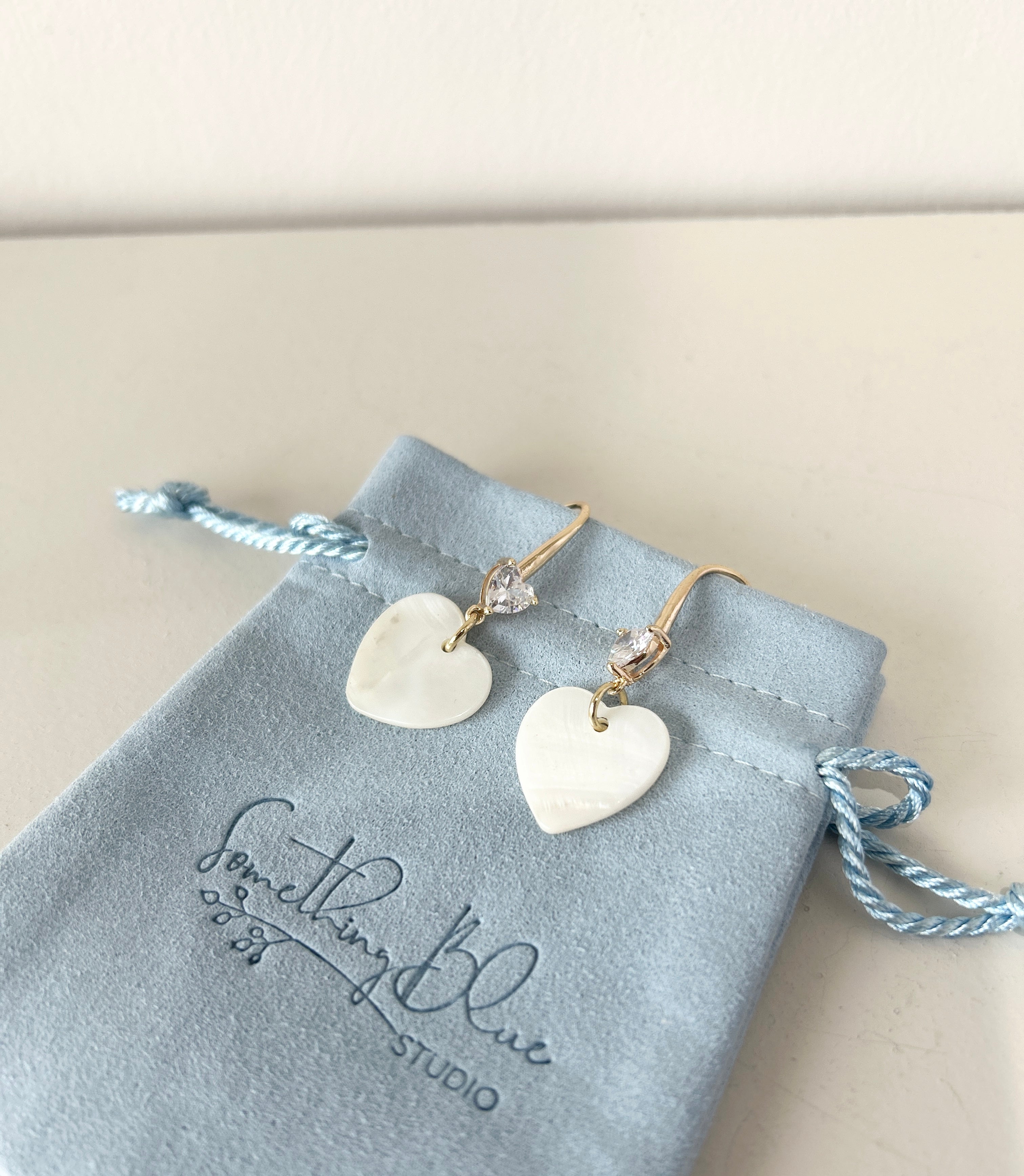 Heart-shaped earrings on a velvet jewellery pouch with 'Something Blue Studio' branding.