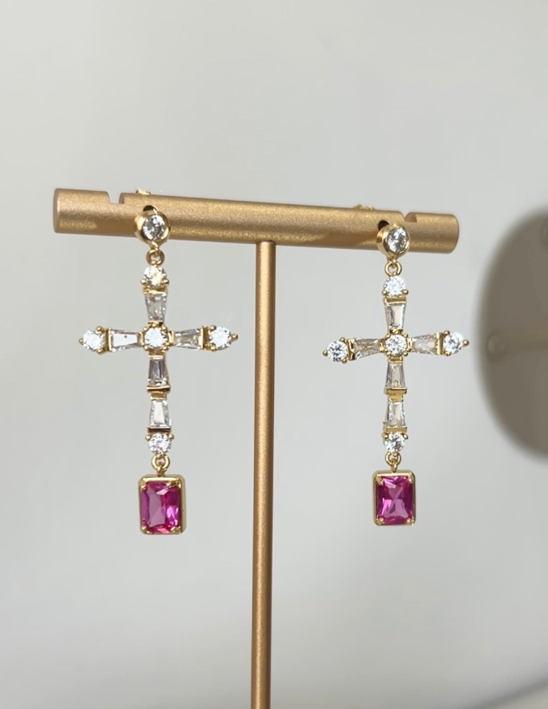 Gold earring stand with pink gemstone cross shape earrings on a white surface.