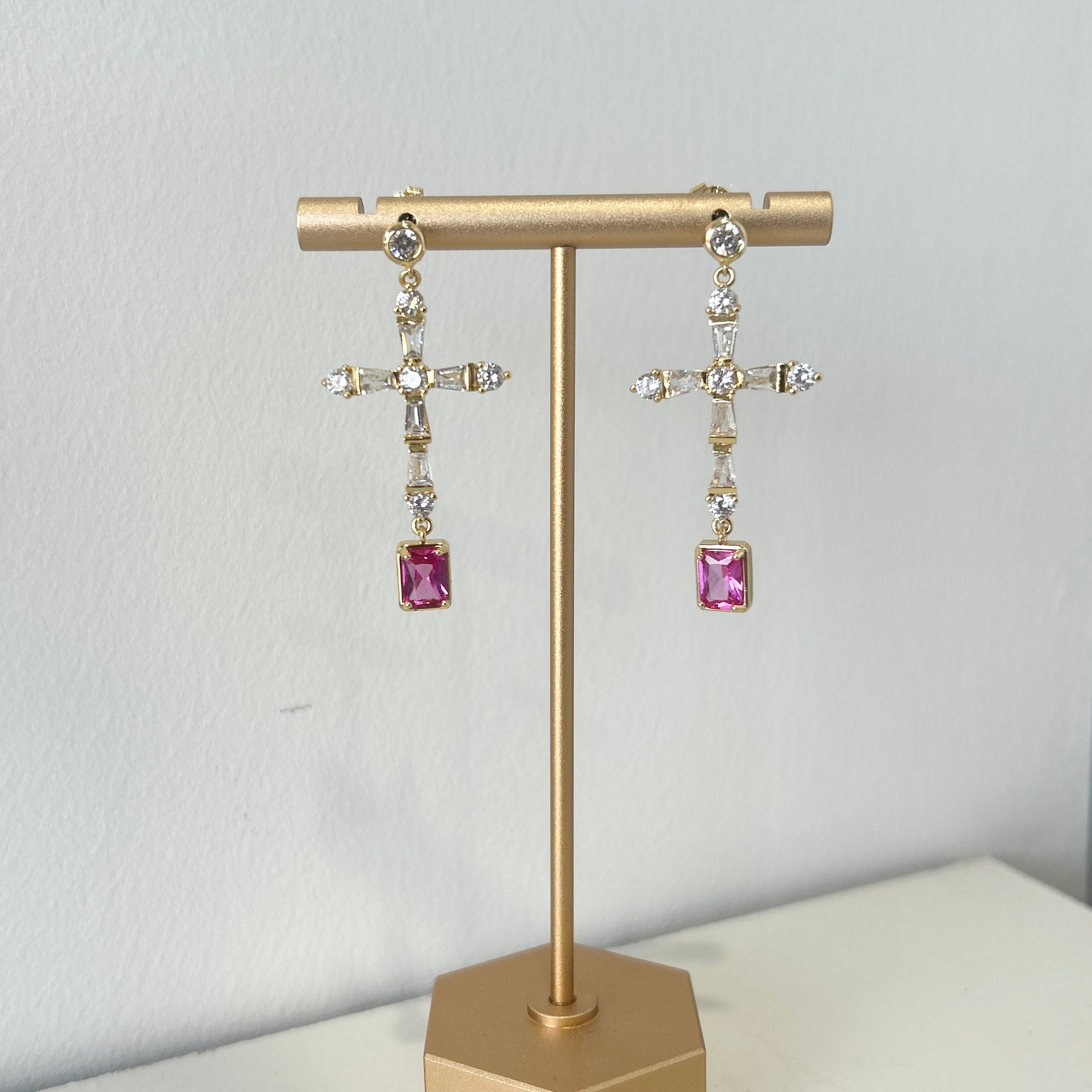Gold earring stand with pink gemstone cross shape earrings on a white surface.