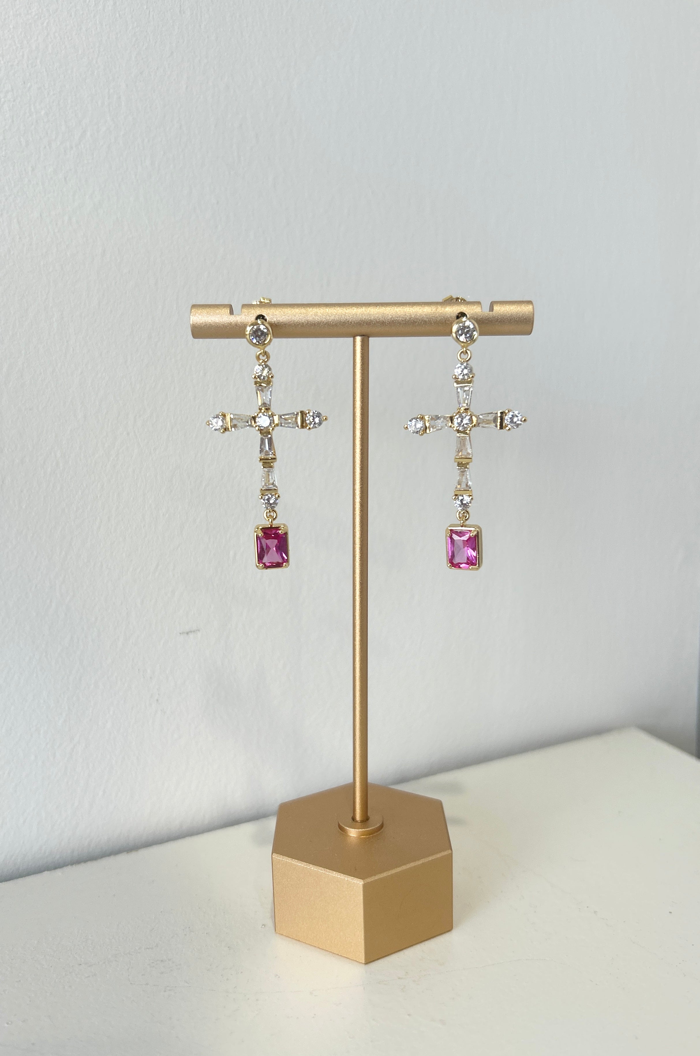 Gold earring stand with pink gemstone cross shape earrings on a white surface.