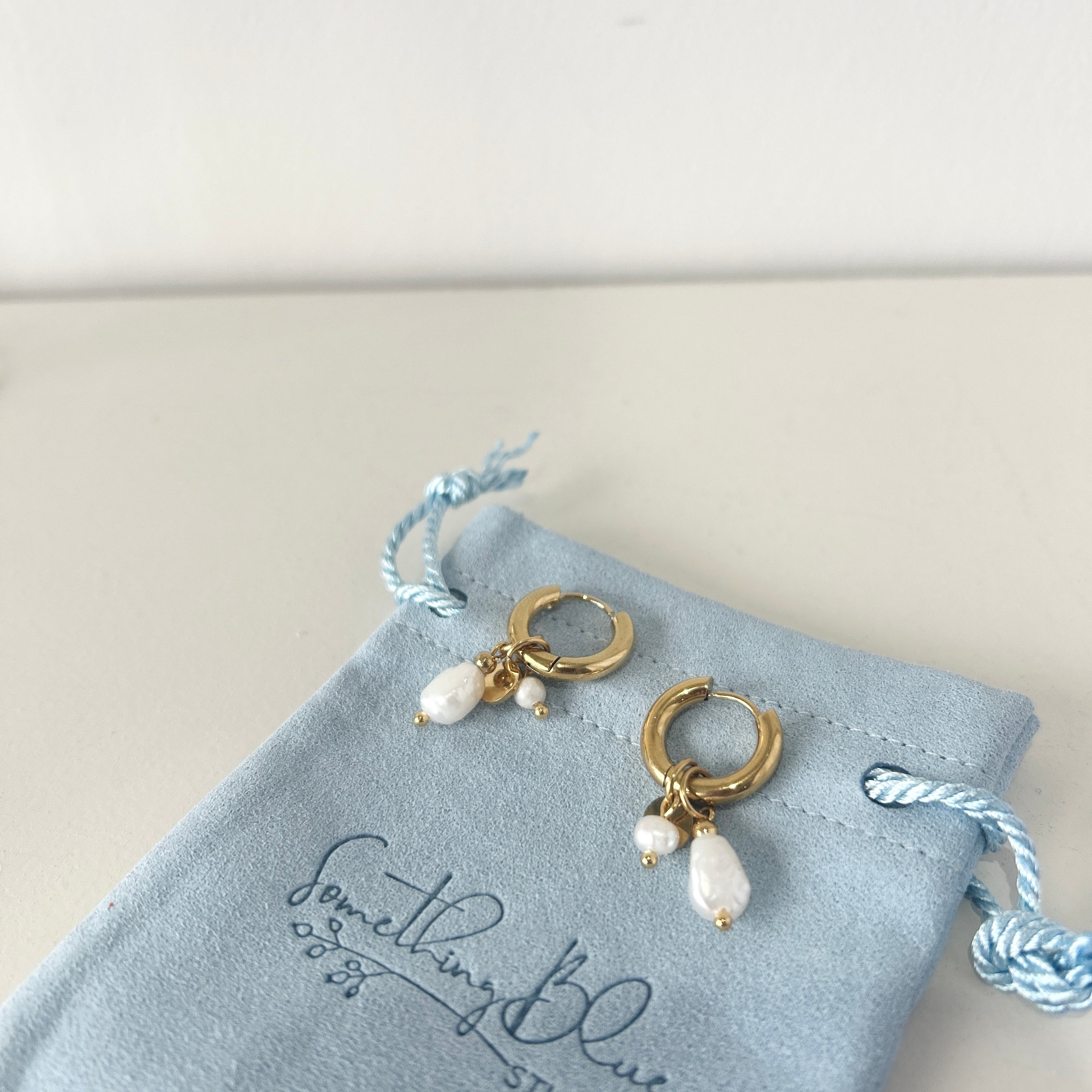 Gold earrings with pearl drops on a 'Something Blue Studio' jewelry pouch.