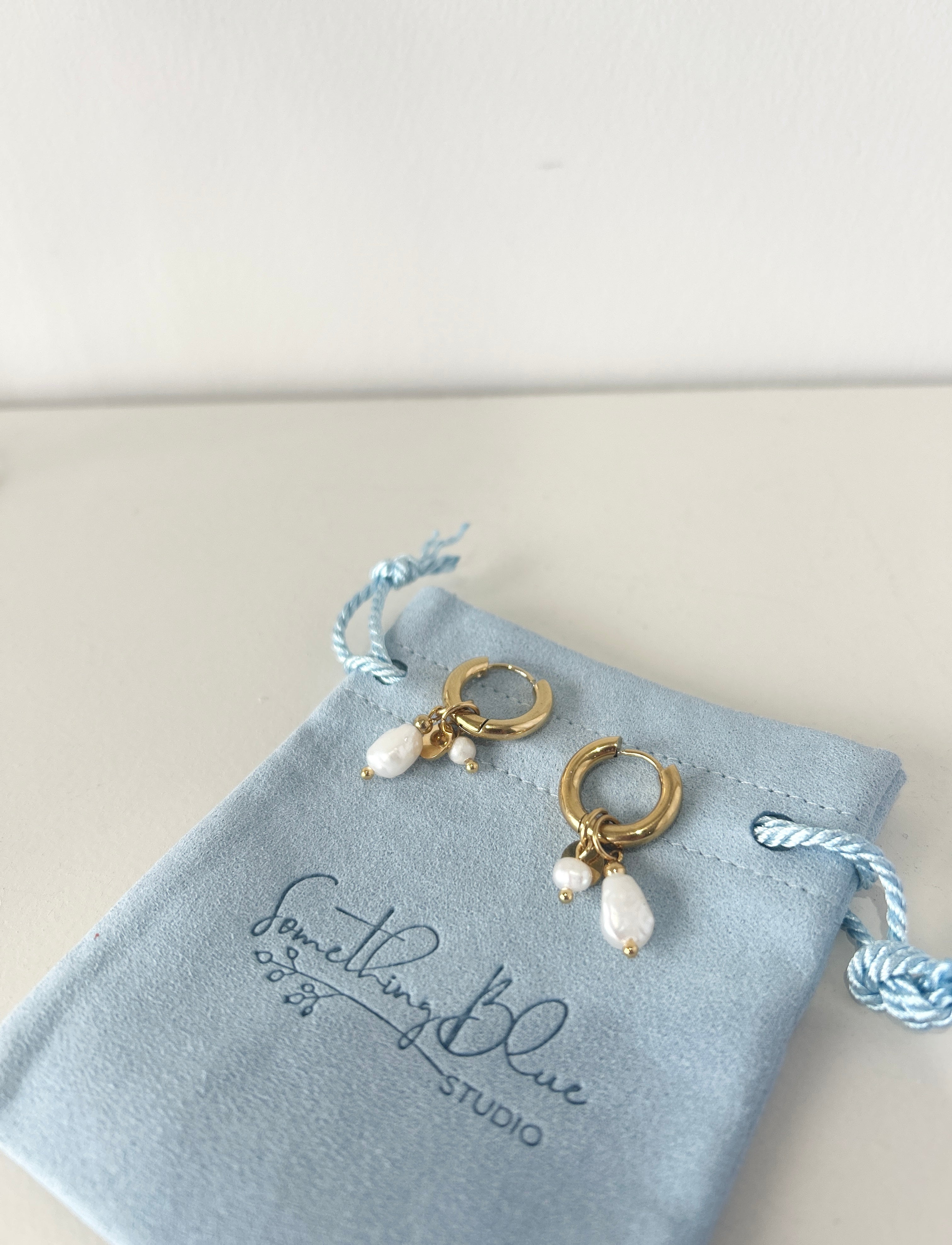 Gold earrings with pearl drops on a 'Something Blue Studio' jewelry pouch.