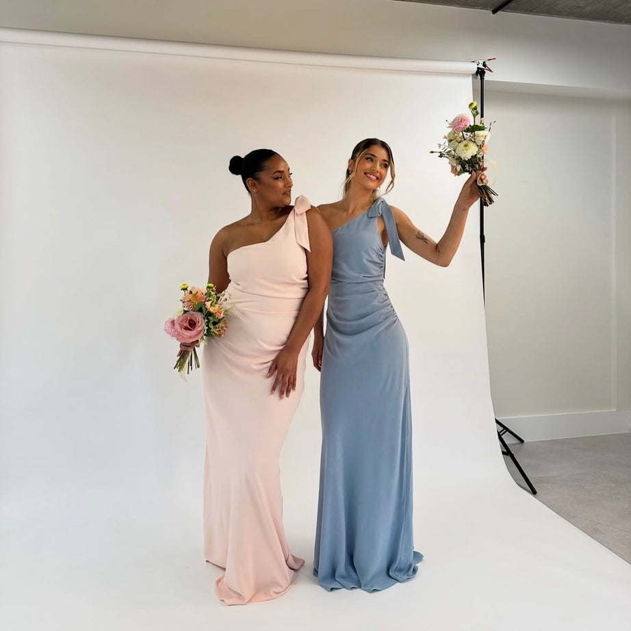 Two women in one-shoulder dresses holding flowers in a studio setting.