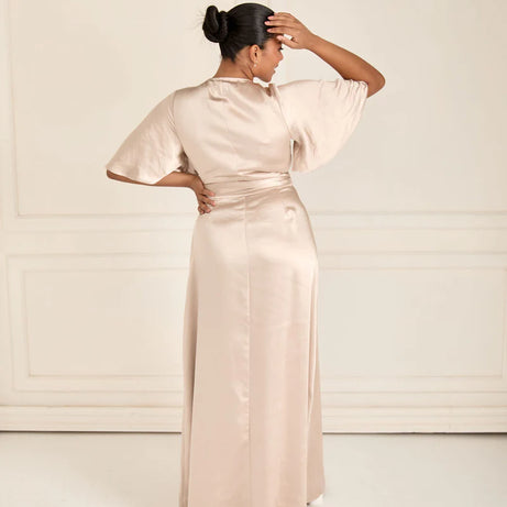 Woman wearing a long, champagne satin wrap bridesmaid dress with a high slit on a white background