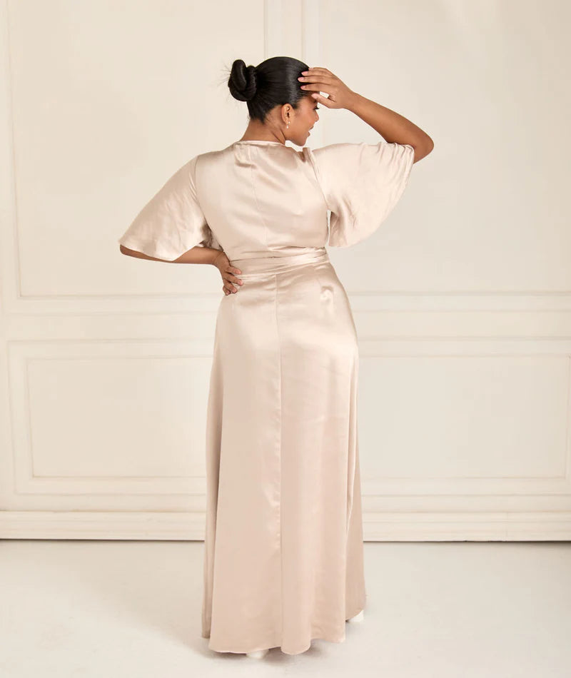 Woman wearing a long, champagne satin wrap bridesmaid dress with a high slit on a white background