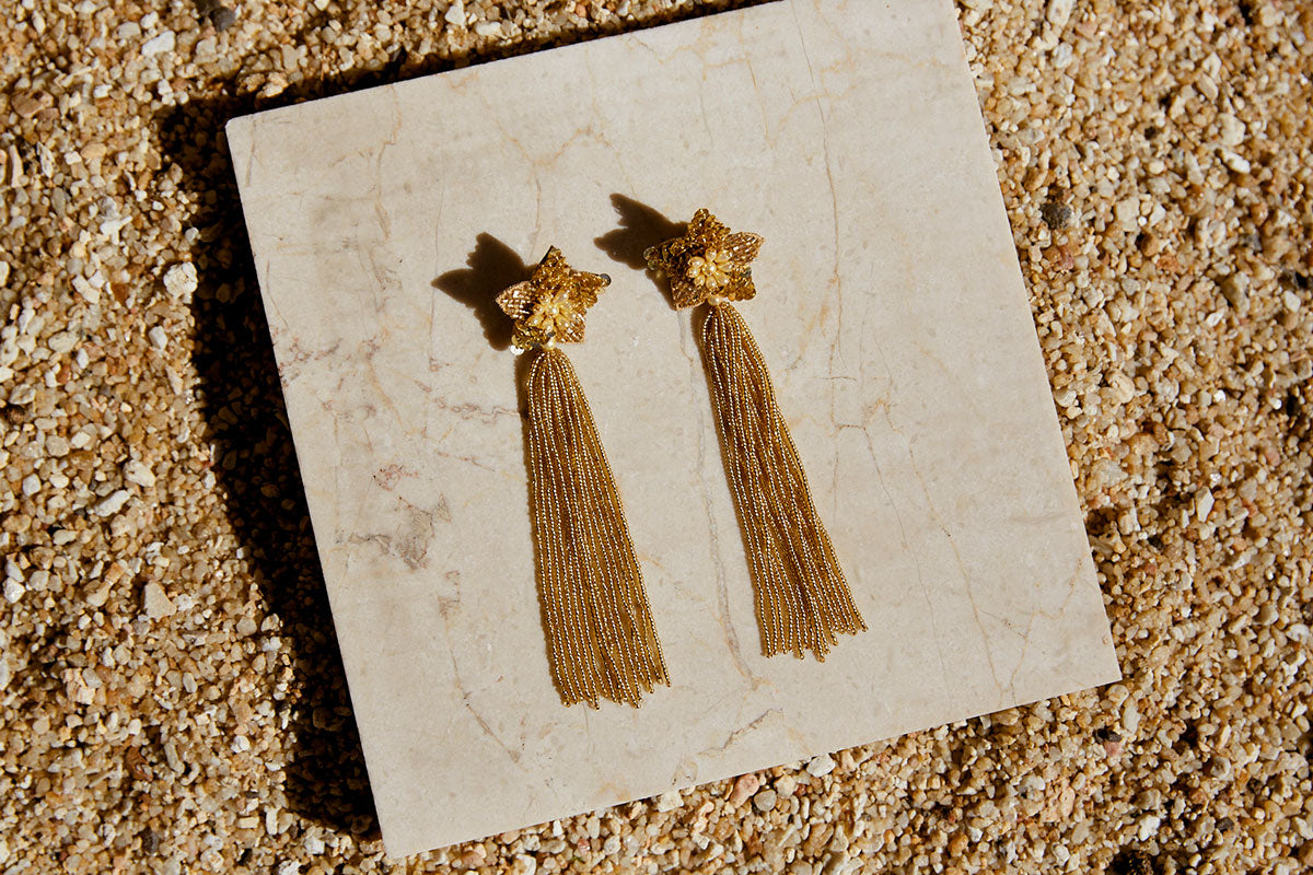 Image of long star shaped gold beaded tassel earrings against a sandstone backdrop.
