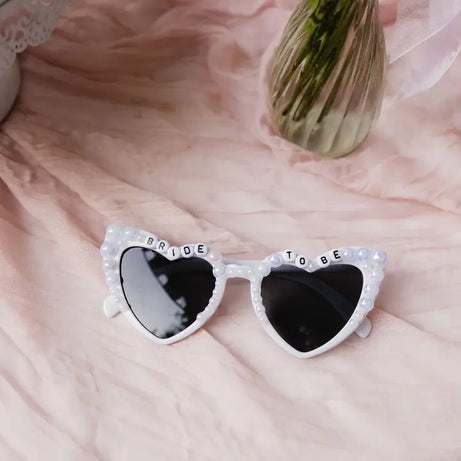 Heart-shaped sunglasses with 'Bride To Be' text on a soft pink background