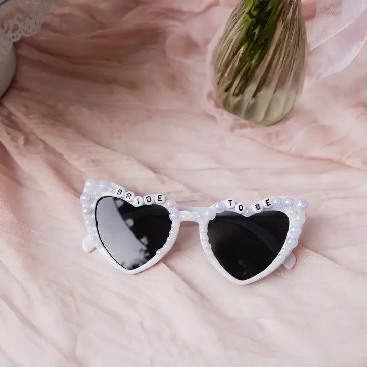 Heart-shaped sunglasses with 'Bride To Be' text on a soft pink background
