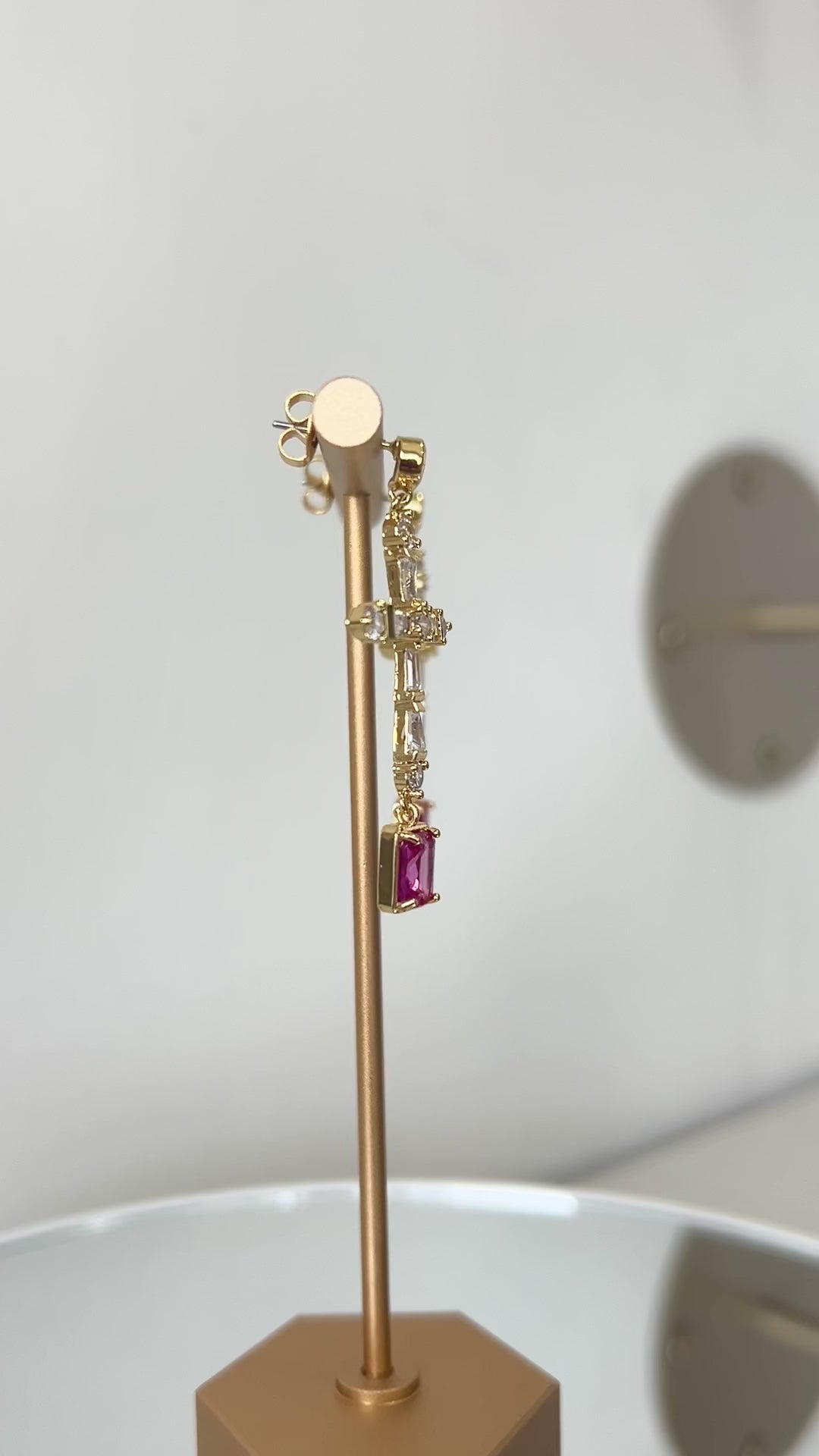 Gold earring stand with pink gemstone cross shape earrings on a white surface.