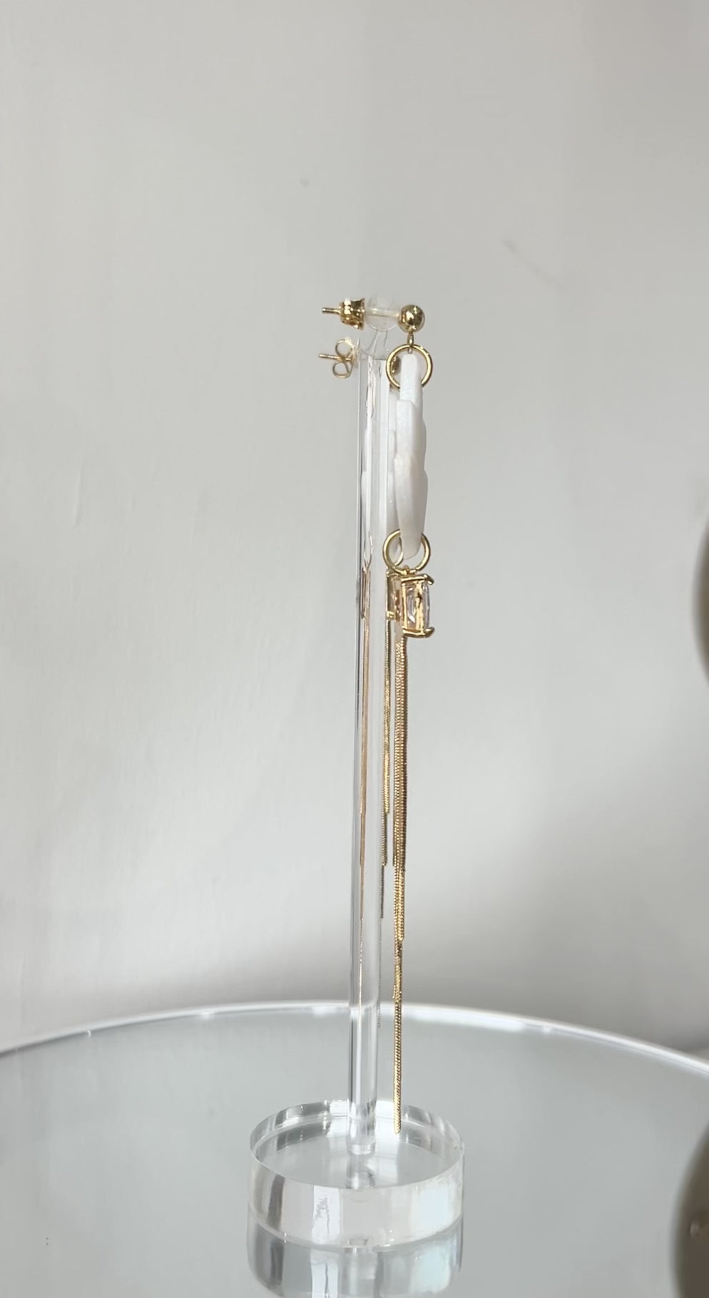 Video clip of statement earrings with white floral elements and gold tassels on a stand against a light gray background.